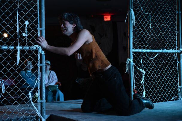 An actor kneeling over in pain, holding onto a chain link fence
